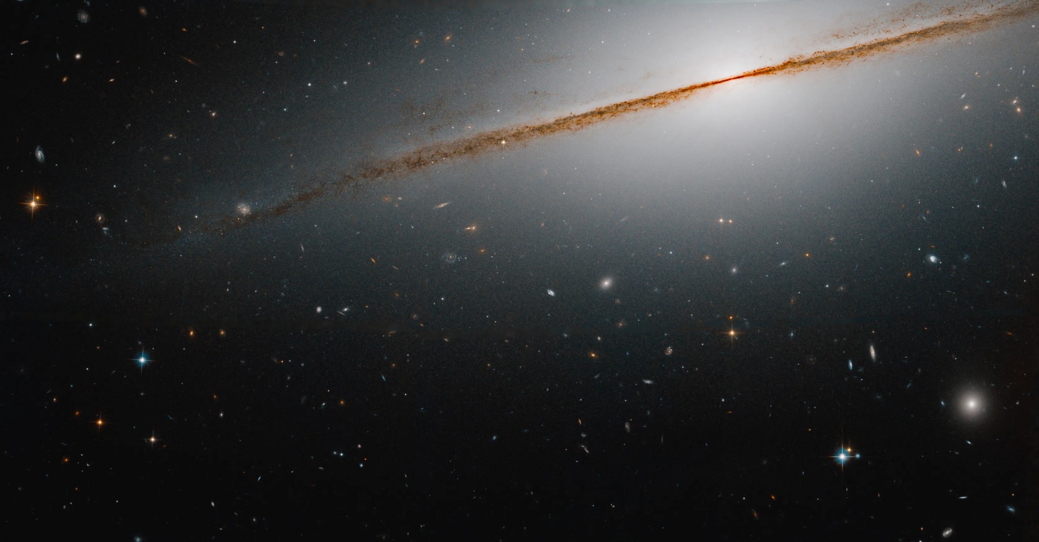 Edge-on galaxy, with a distinctive dust lane, extending from upper right to middle-left of the image. distant galaxies dot the scene.