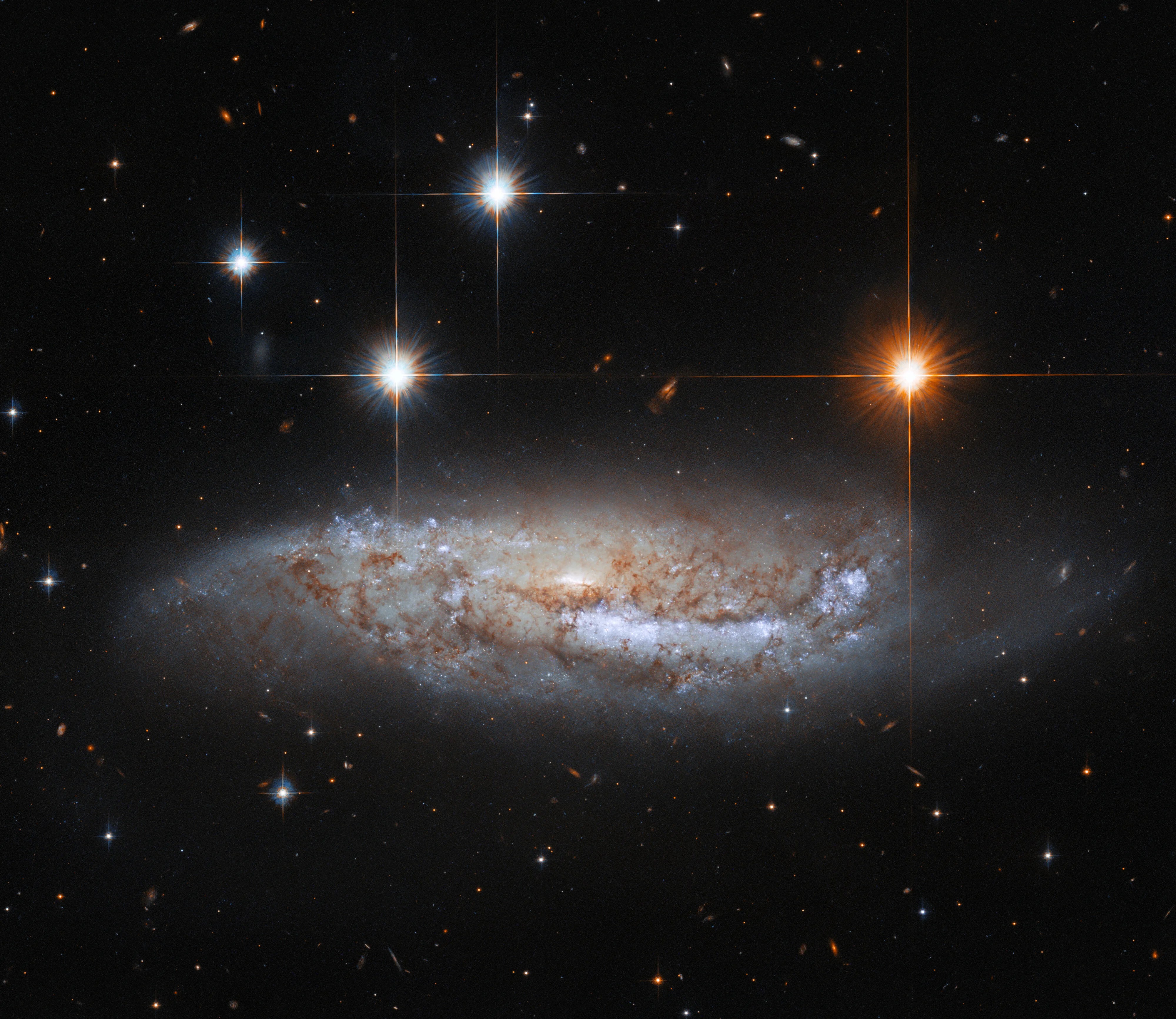 Barred spiral galaxy seen from the side. 3 bright-white foreground stars are to the upper left of the galaxy, one bright-orange-white star is to the upper right.