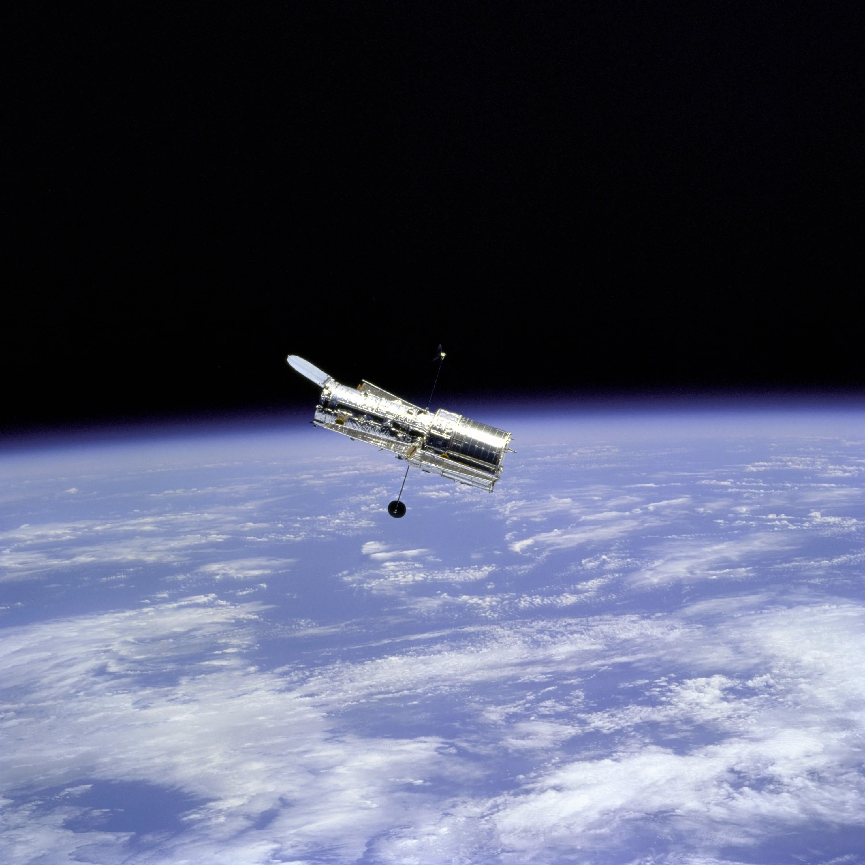Hubble floats against the black background of space with the curve of the Earth below it. Pale blue oceans and white clouds are visible on Earth.
