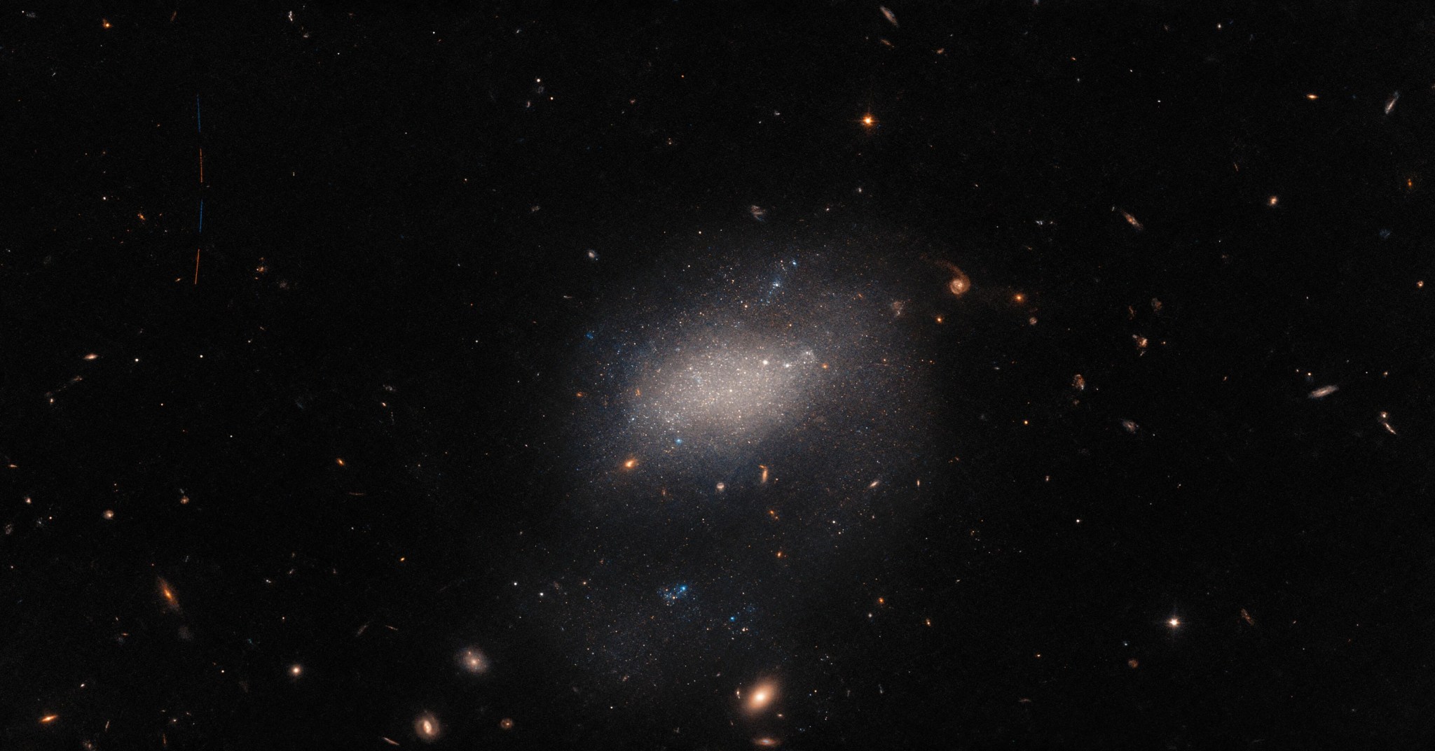 Black field with with galaxies. center of image holds a diffuse collection of stars, the galaxy ugc 7983. upper left: four distinct vertical streaks, alternating rust-red and blue, form a dashed line. the signature of the photobombing asteroid.