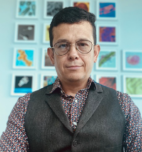 Portrait photo of a man with short brown hair and eye glasses wearing a suit vest over a patterned shirt; several colorful satellite images are on the wall behind him.