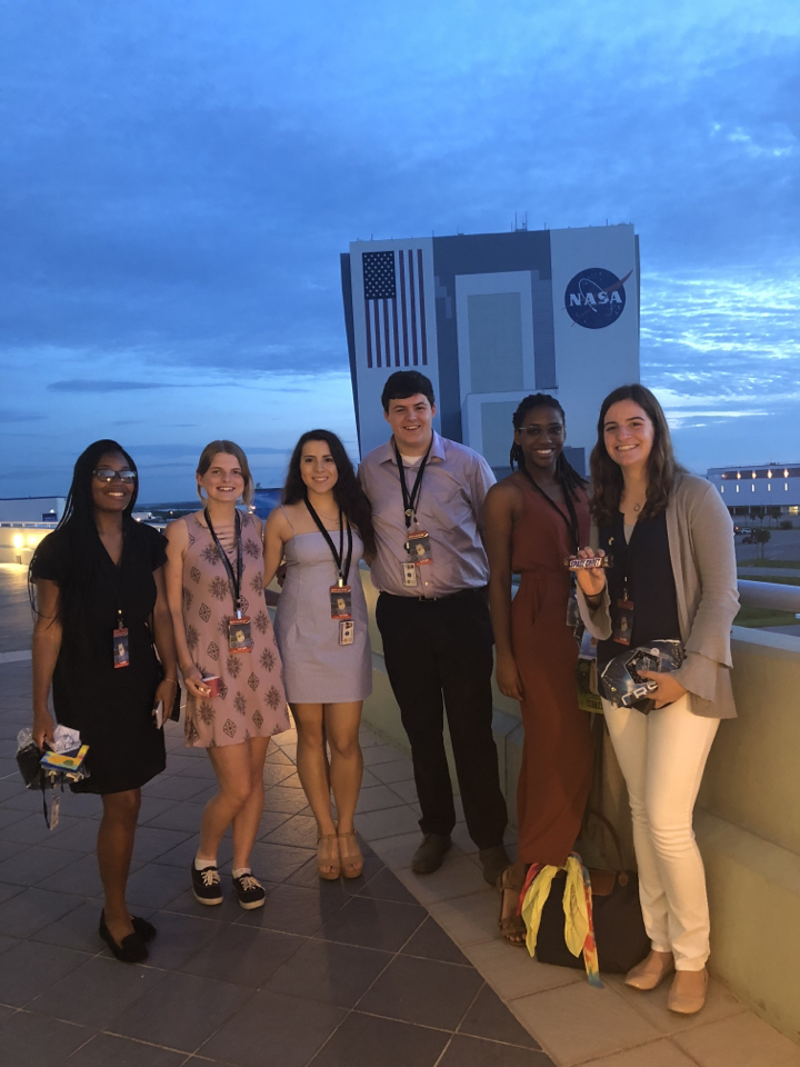 Six young adults standing for a posed photo on a balcony at KSC overlooking the VAB