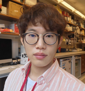 Portrait photo of a young man with short wavy brown hair wearing glasses and a light red white-striped shirt standing in a lab with shelves behind him