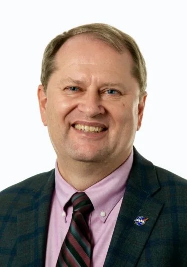 A formal portrait of a smiling caucasian male with blond hair and blue eyes. He is wearing a pink shirt with a striped tie and a dark suit. A NASA lapel is located on his left lapel.