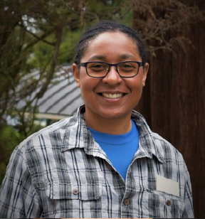 Portrait photo of a dark skinned smiling woman with braided hair, eye glasses and a flannel shirt; trees and a roofline are in the background.