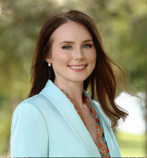 Portrait photo of a smiling woman with long red-brown hair wearing a white coat and earrings, blurred trees are in the background
