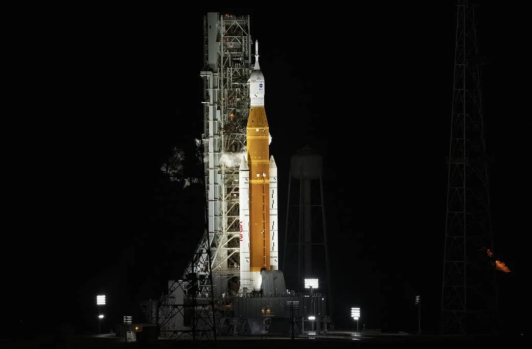 高耸的阿耳特弥斯一号火箭在黑色背景下发光。火箭底部周围的发射台灯光照亮了火箭。带有亮橙色和白色机身的火箭支架位于灰色发射架的前面。