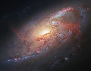 A bright yellow galaxy core, surrounded by spiral arms with pink and brown lanes of dust and bright regions of star formation.