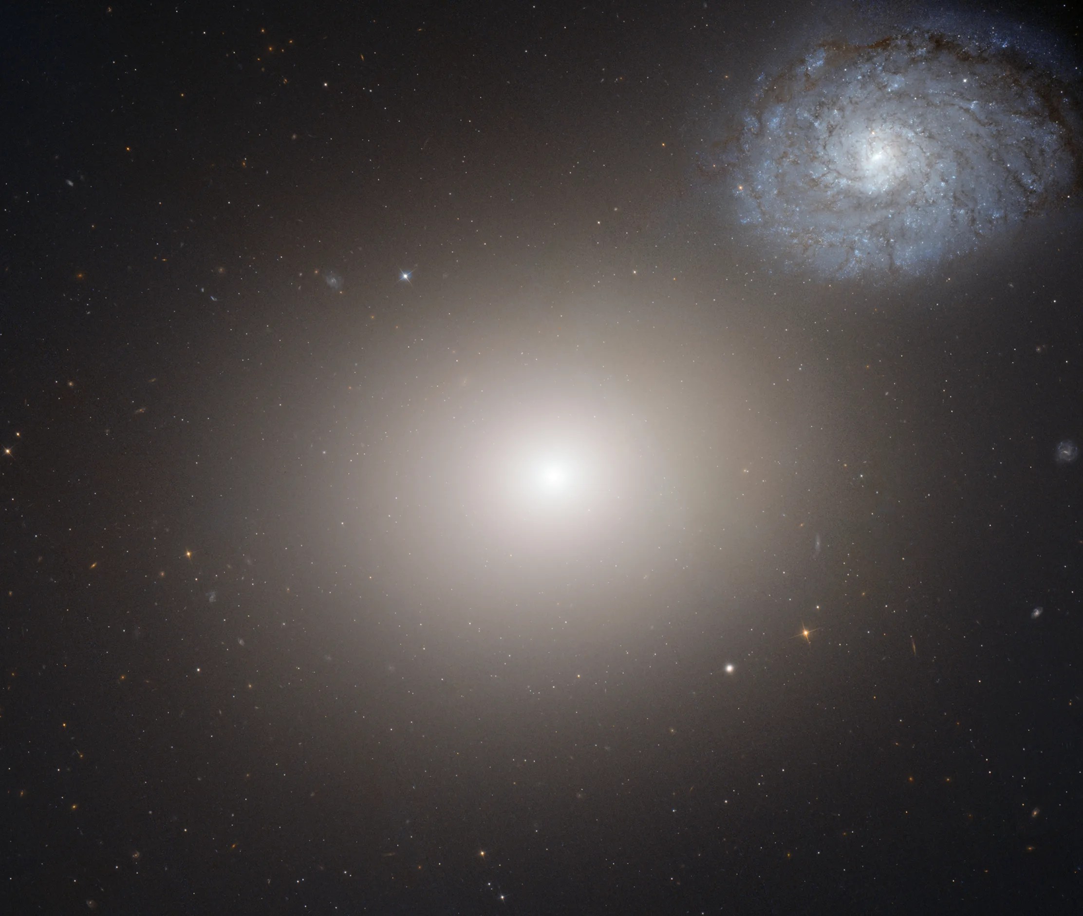 A hazy white-yellow light shines in the center of the image against black space, dotted with faint, distant stars. In the top right of the image is a spiral galaxy with many arms as seen from overhead. It is light blue.