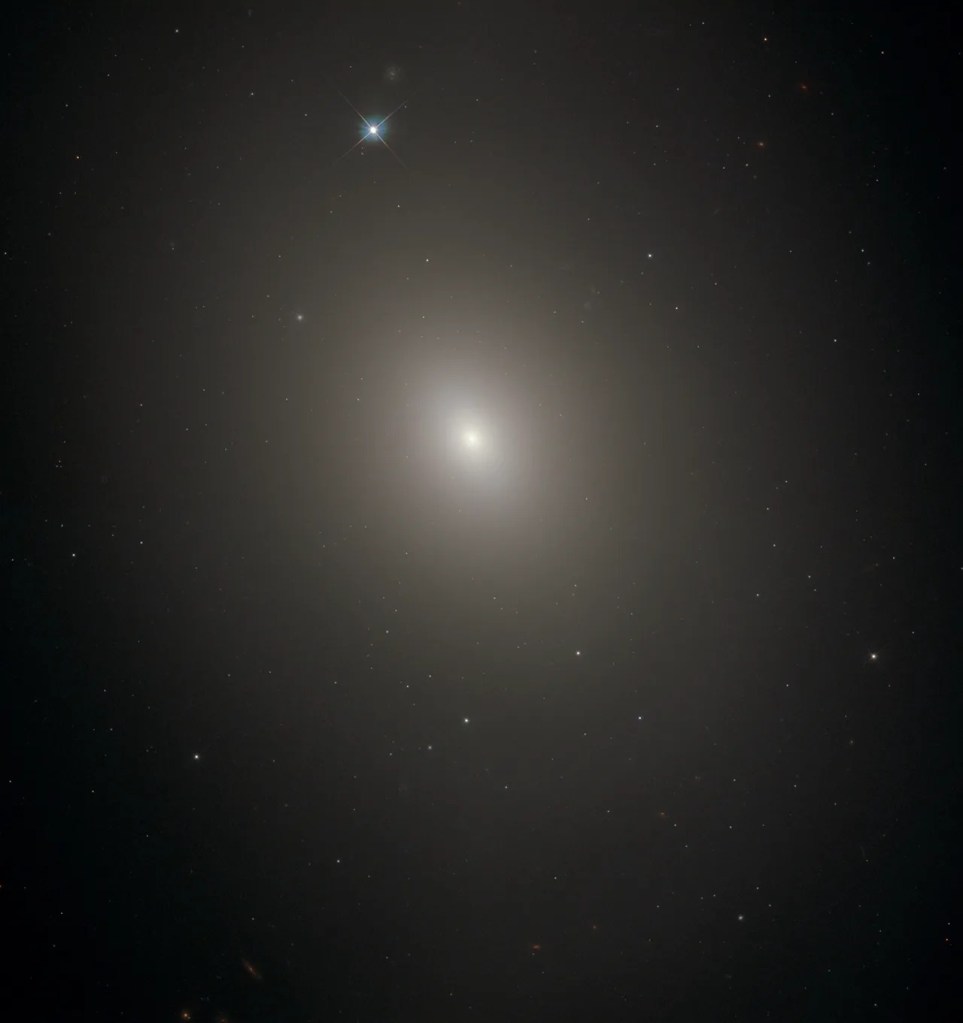 Diffuse oval haze against black background. Stars and distant galaxies shine through it. Just above image center the haze gets brighter and denser toward the bright-white core of the galaxy. Top, just left of center is a bright foreground star.