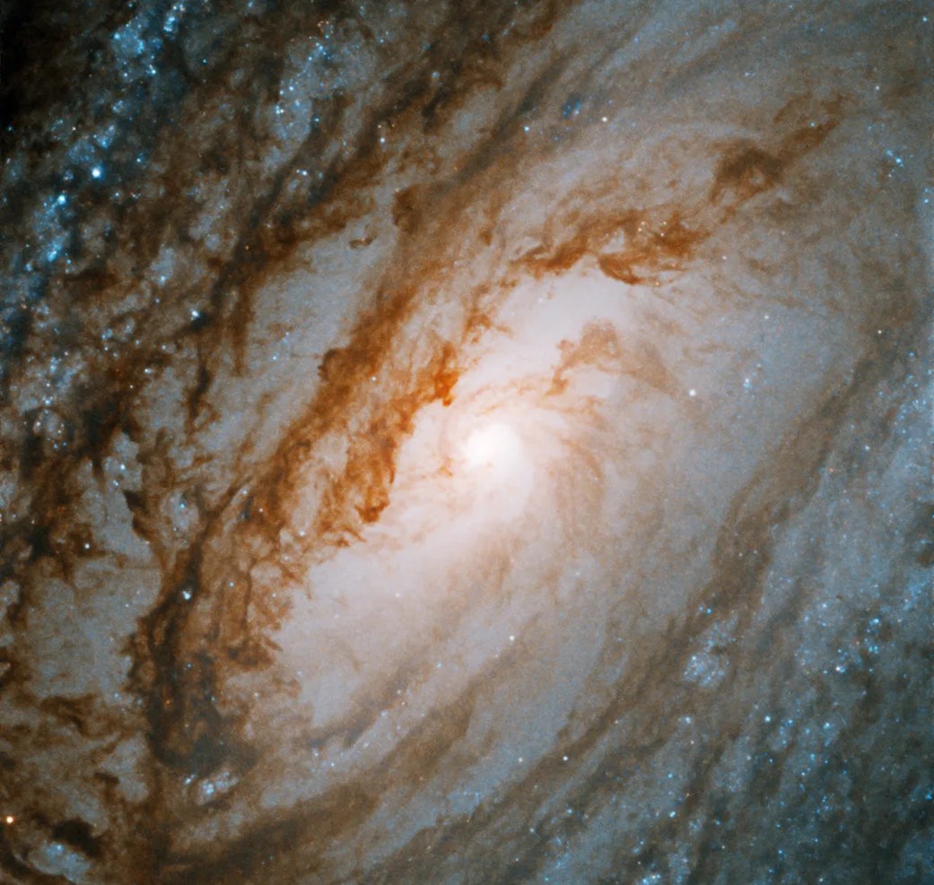 A bright galaxy core shines at the center of the image, surrounded by large spiral arms laced through with dark dust and bright blue regions of star formation.