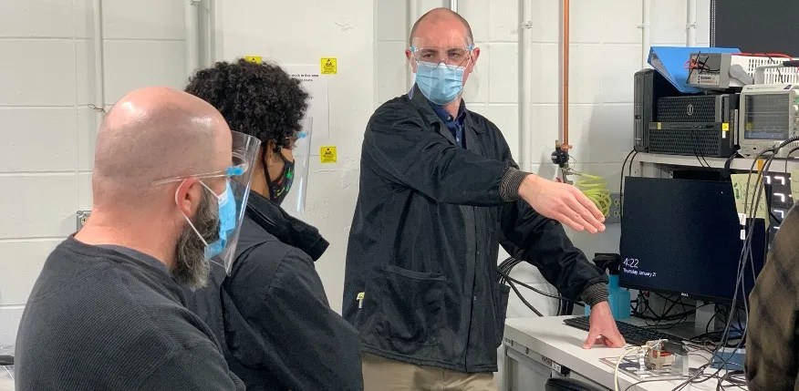 Photo of three men in a lab wearing dark lab jackets and face masks; one man is standing and pointing at the equipment while looking at the other two seated men.