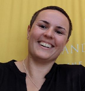 Portrait photo of a smiling woman with short brown hair, wearing a dark shirt and necklace in front of a yellow backdrop.