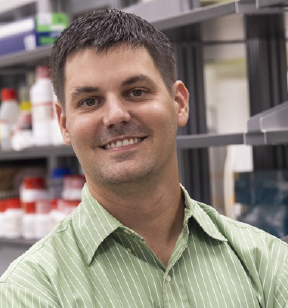 Portrait photo of a smiling man with short dark brown hair and a light green and white striped button down shirt standing in front of shelves in a lab stocked with supplies