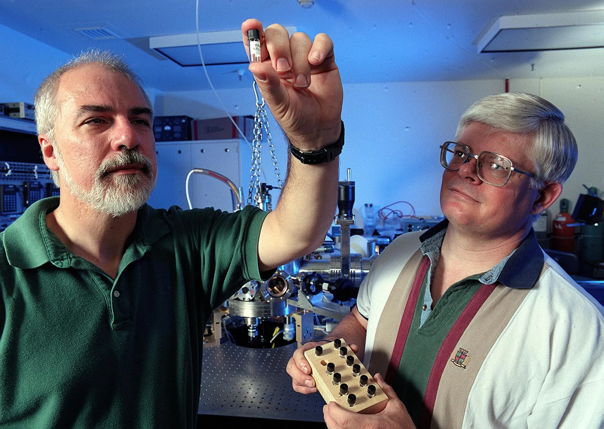 Two men standing in the foreground. The man on the left is holding up a small glass vial, while the man on the right holds a small tray of vials (one spot is unoccupied; it is being held by the man on the left).