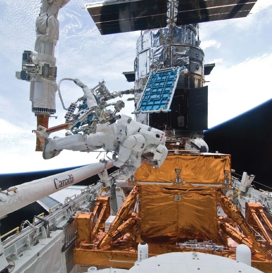 Astronaut Mike Good in his spacesuit with his feet attached to the shuttle's robotic arm works on Hubble against a backdrop of Earth and space.