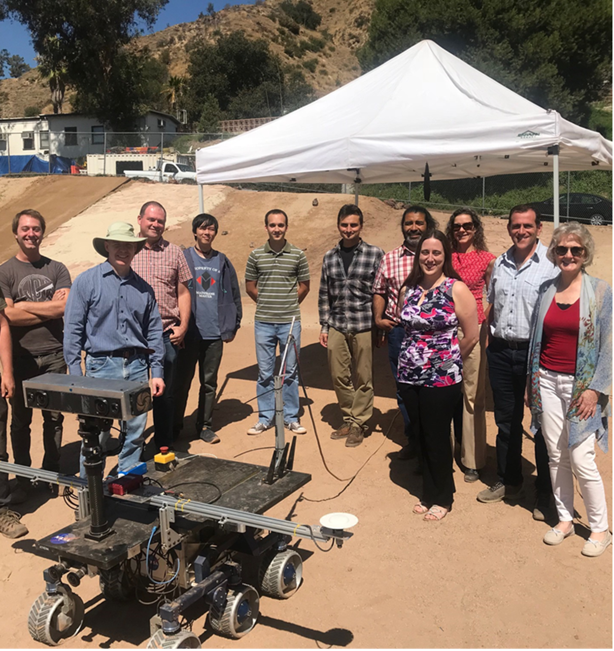 Photo of group of engineers standing on soil with a model of a rover