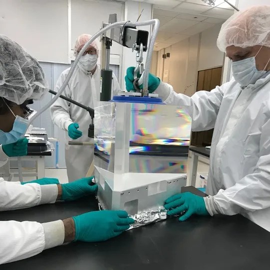 Photo of three lab scientists in white lab gear wearing turquoise gloves and installing a prism into a white metal piece of equipment