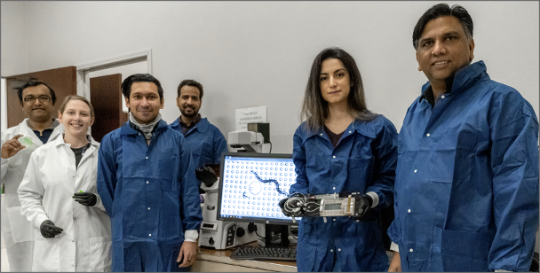 Group photo of 6 research scientists in blue and white lab gear holding lab equipment.