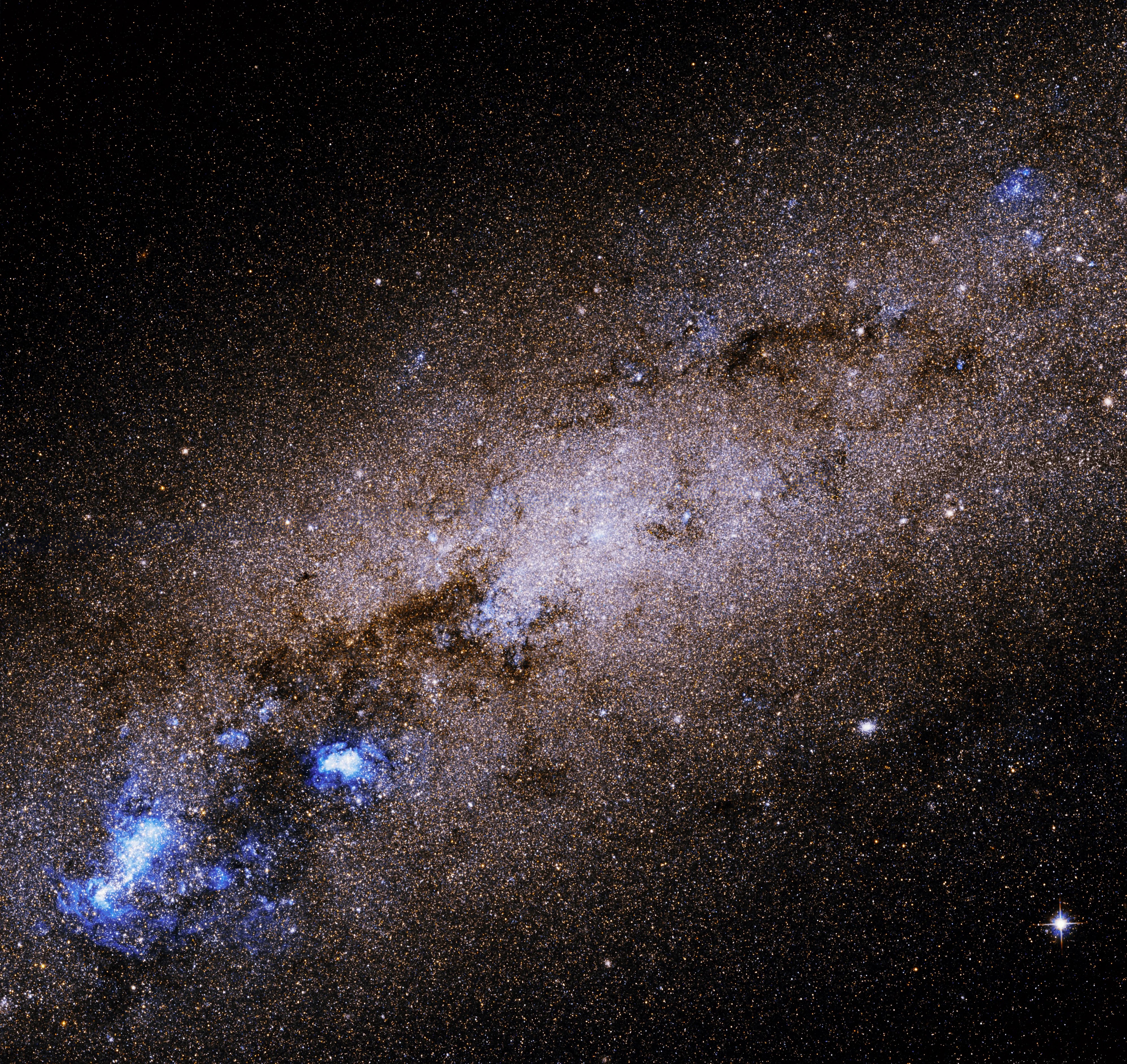 The center of the galaxy glows in a pinkish-white, and is tilted diagonally, going up-right and down-left. Black clouds of dust are visible around the center, and in the lower left corner, thick blue "clumps" of stars.