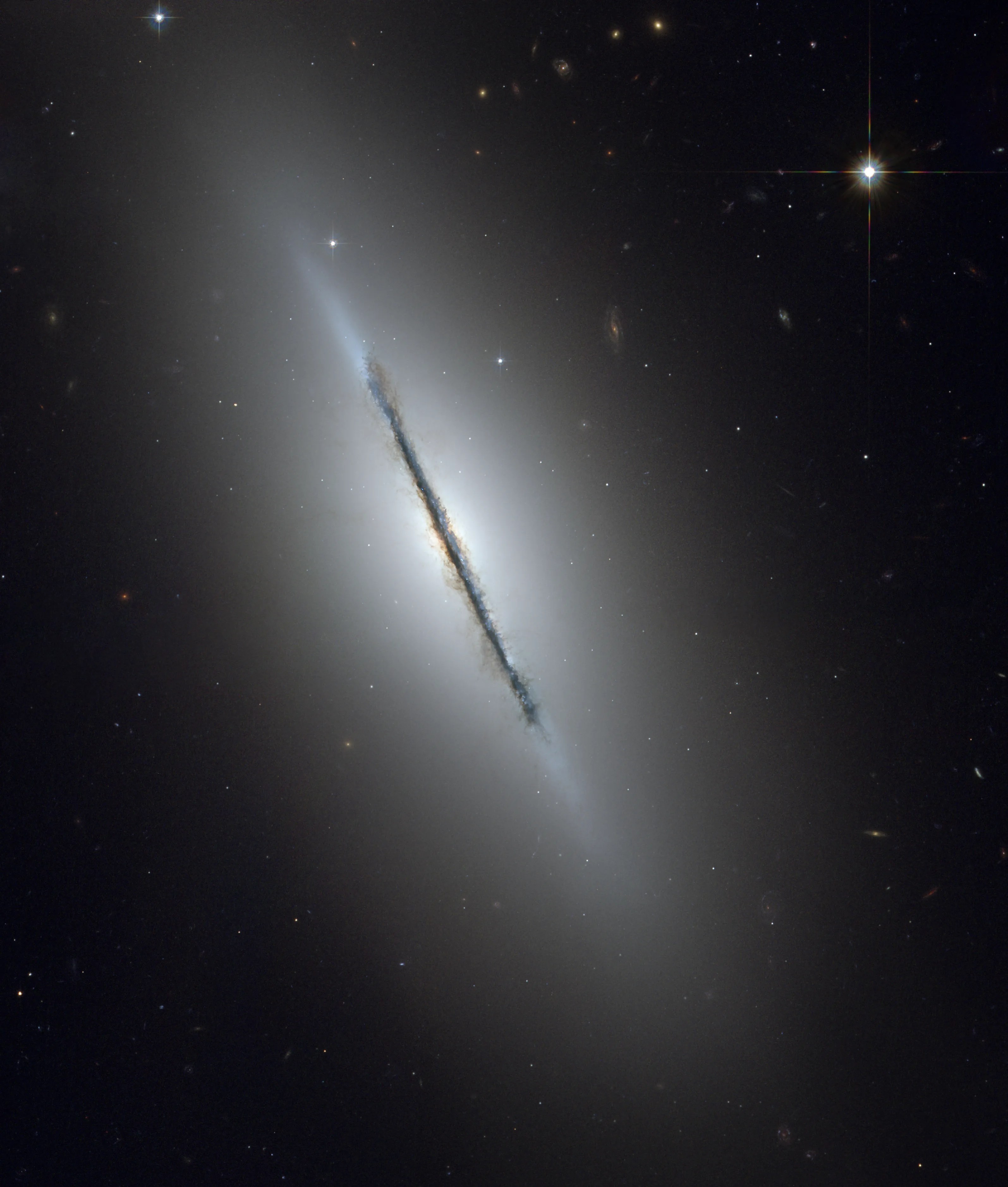 A galaxy is seen edge-on, with a dark lane of dust obscuring it surrounded by a hazy white glow, all against a black background.