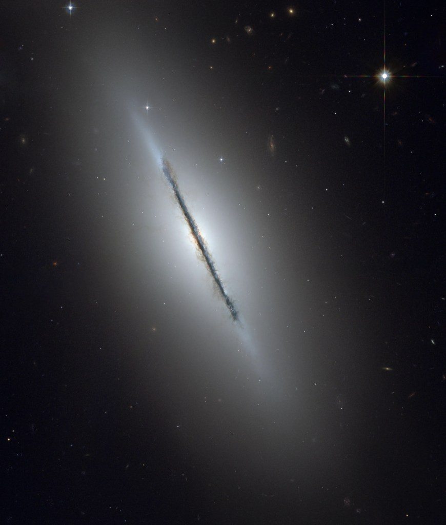 A galaxy is seen edge-on, with a dark lane of dust obscuring it surrounded by a hazy white glow, all against a black background.