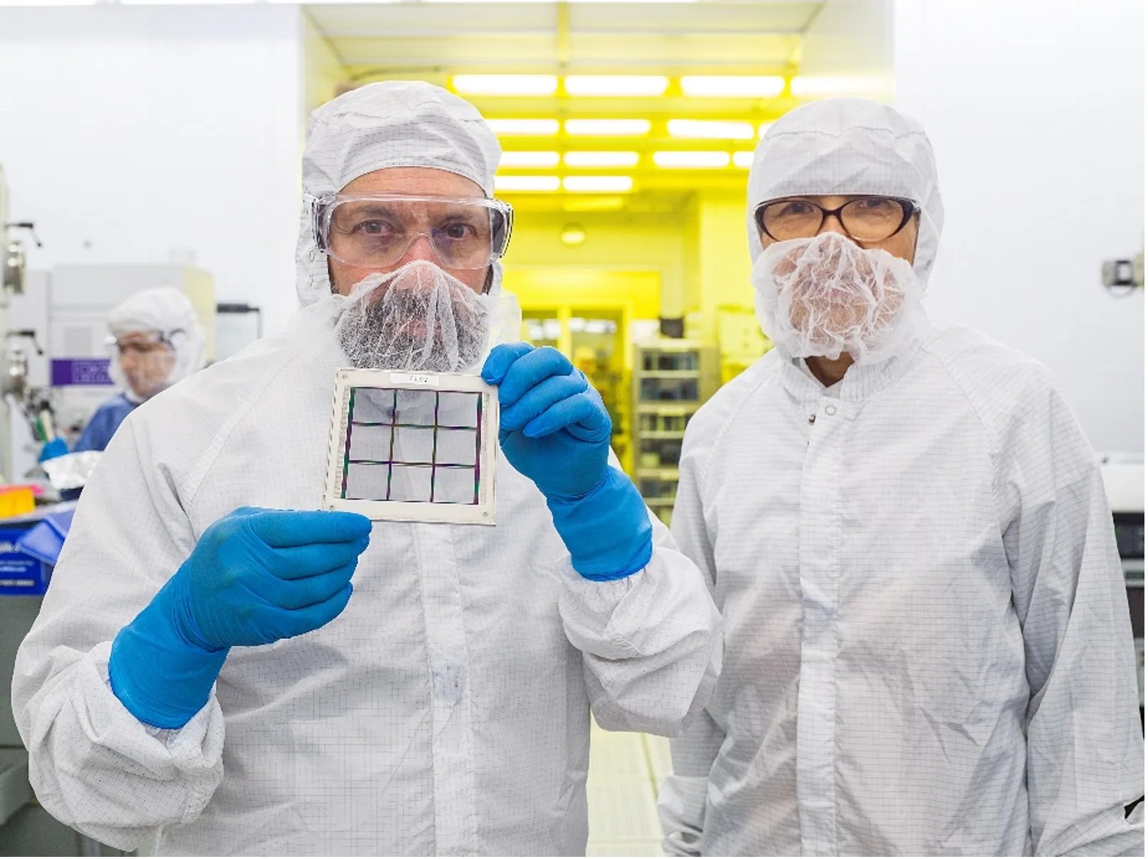 Two scientists wearing white lab jumpers in a lab; the male on the left is wearing blue rubber gloves and holding an array prototype that looks like a small grid.