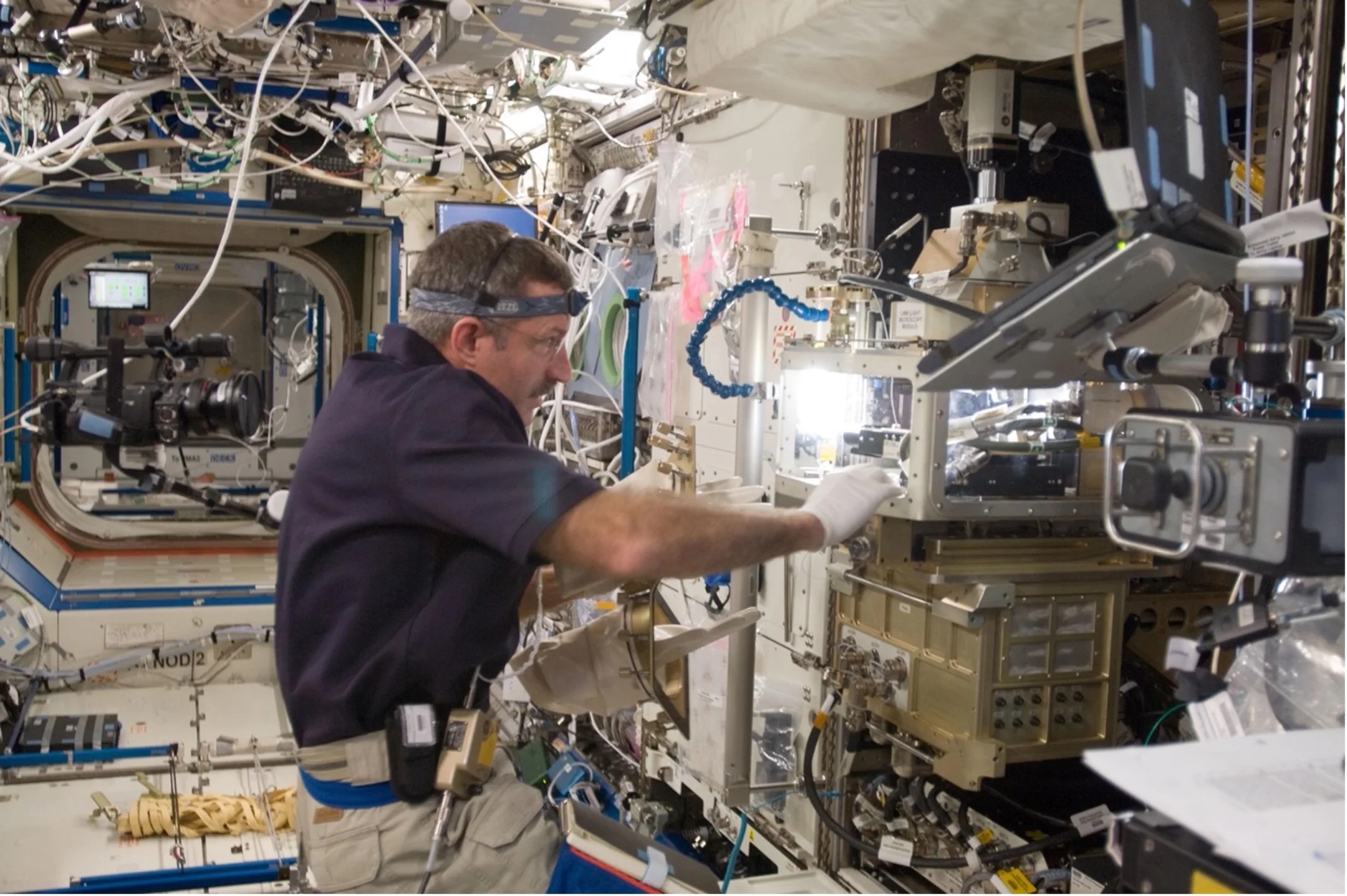 Astronaut aboard the International Space Station holding onto large, sqaure opening within a mechanical device.