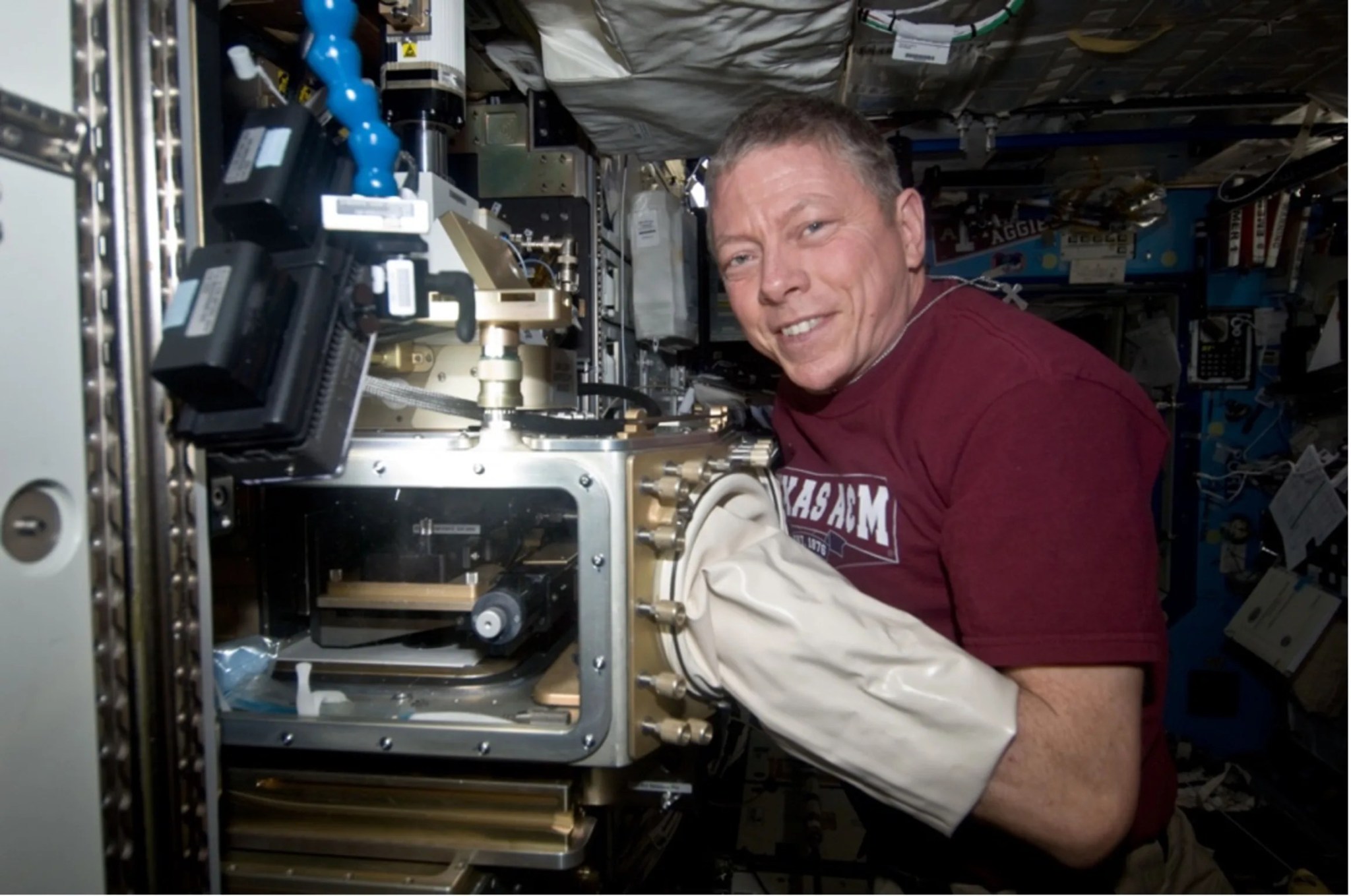 Astronaut with his hand inside a large, white latex glove attached to a gold, mechanical device.