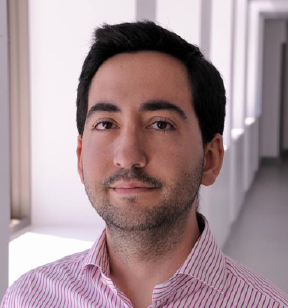 Portrait photo of a young man with short dark hair and facial stubble wearing a red and white striped shirt and standing in a hallway with natural light beaming in