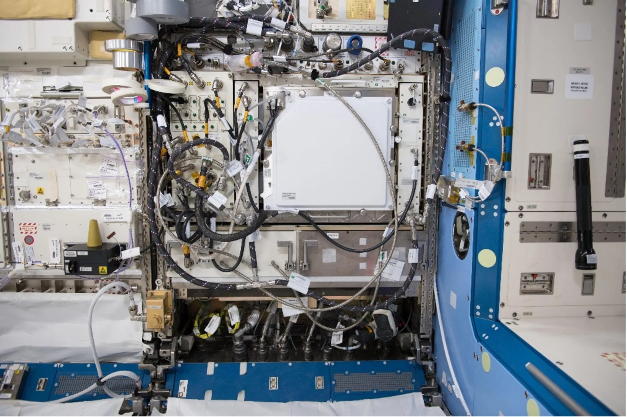 A complex metal box is set into the inside wall of the space station. Black and silver wires and tubes crisscross the front surface of the box, connecting the environmental control system in the left half of the box to the plant growth chamber in the right half.