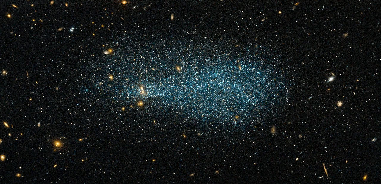 An asymmetrical blob of bluish stars on black with golden galaxies