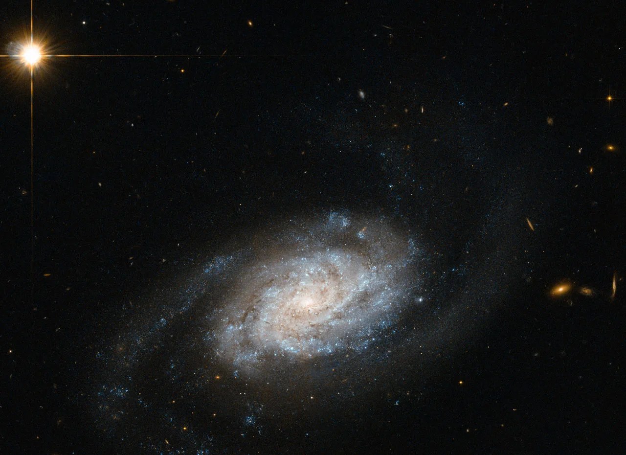 A spiral galaxy with blue-white arms and a hazy orange glow. Distant orange galaxies can be seen in the background off to the right.