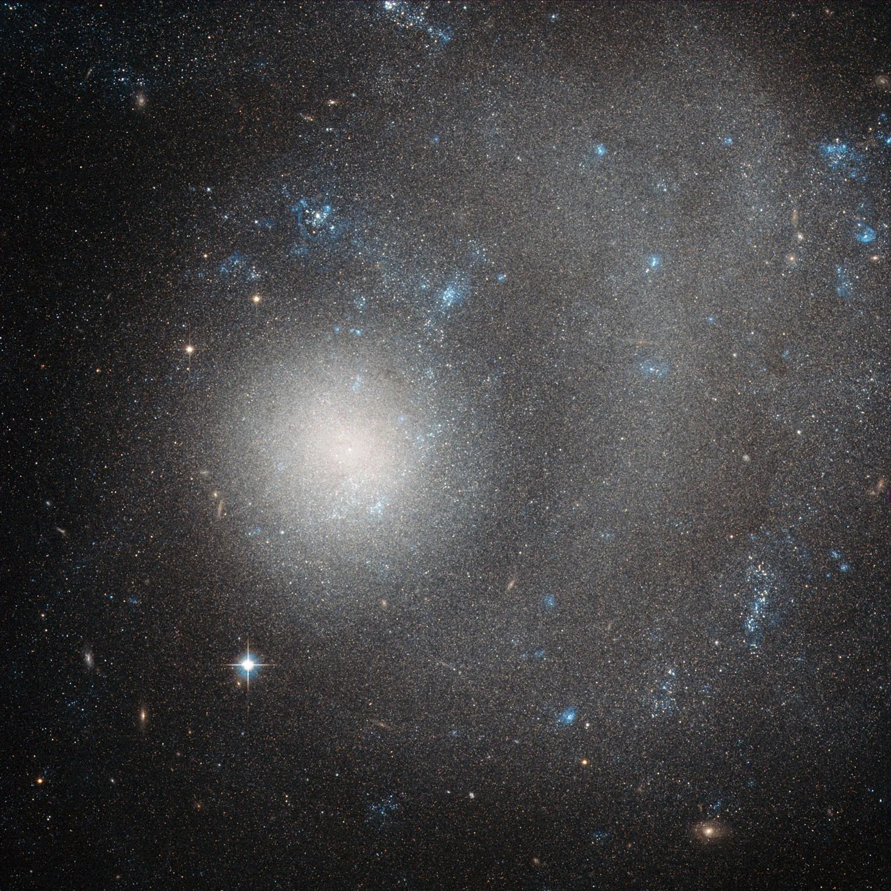 Hubble image of dwarf galaxy ngc 5474;  a diffuse blob of stars off-centered with a vague swirl stretching right and up.