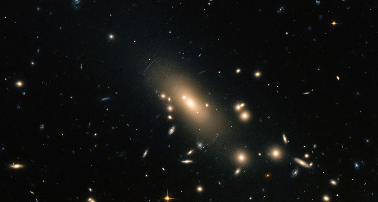 A collection of galaxies glowing in a soft golden light. In the center is an elliptical galaxy that appears to be emanating a bar of golden light. Around it are scattered multiple smaller galaxies of varying sizes, shapes, and colors.