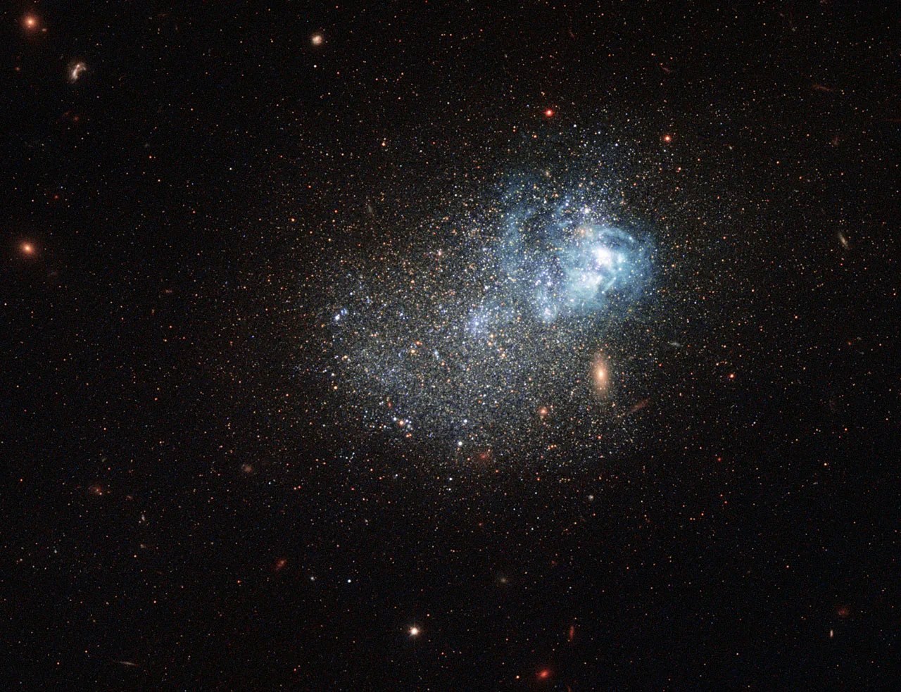 An irregular but somewhat spherical collection of stars, with a large bluish knot of gas visible to the left side of the grouping. Other farther galaxies are visible in the background.