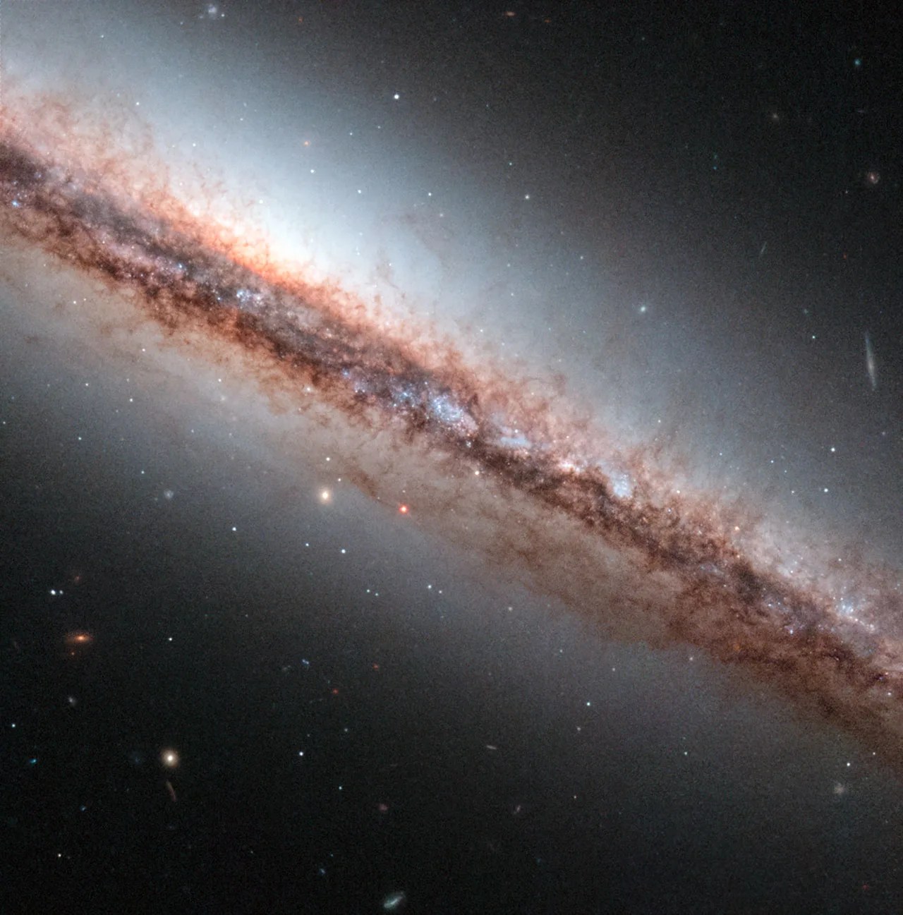 Edge on spiral galaxy with dark dust clouds and a surrounding hazy glow slashes across the frame from the upper left corner to the lower right corner.