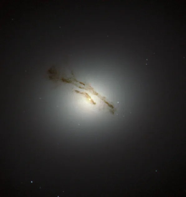 A hazy, yellow-white galactic core shines against black space, with a couple ribbons of dark dust in front of it.