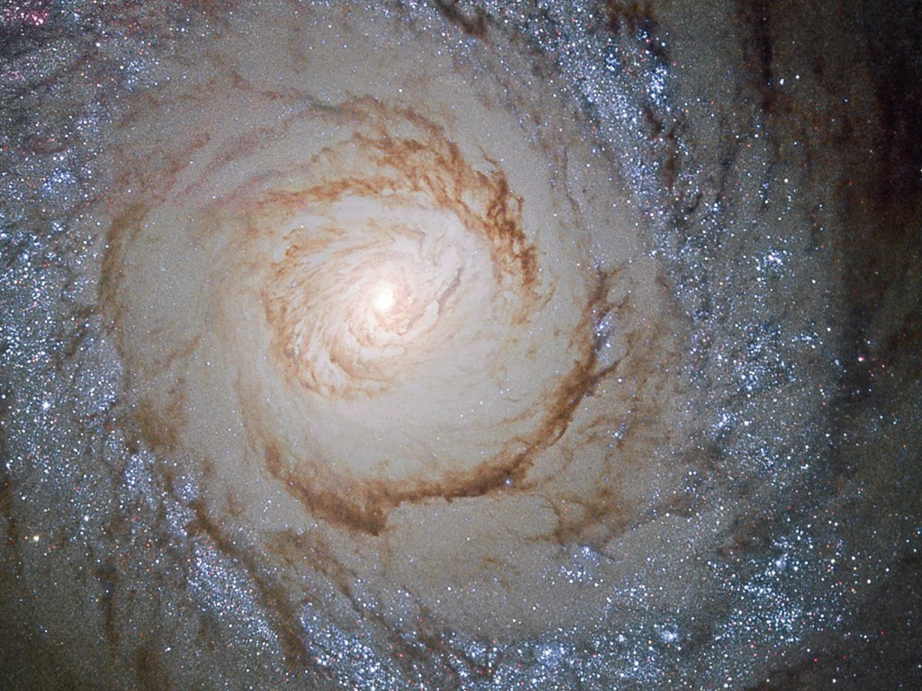 A bright yellow galaxy core shines, surrounded by spiraling arms laced through with dark dust and blue-white regions of new stars.