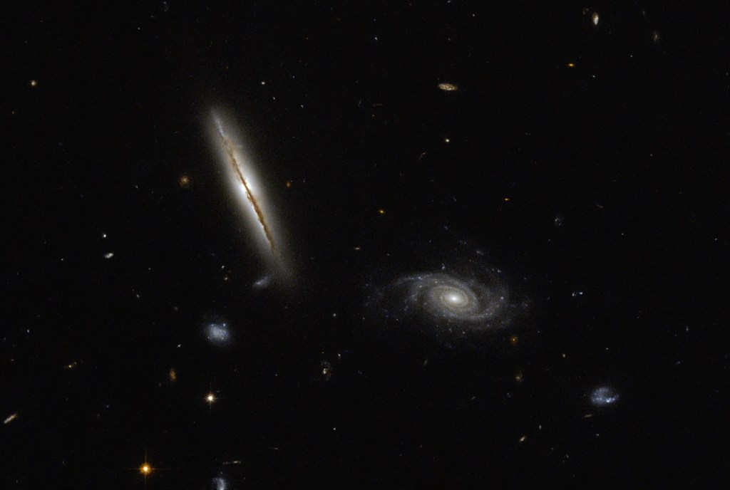 Two spiral galaxies: the main one on the left is seen nearly head-on, just able to see the brownish dust of the disk with a yellowish glow. Its companion spiral is seen more face-on, with grayish-yellow spiral arms.
