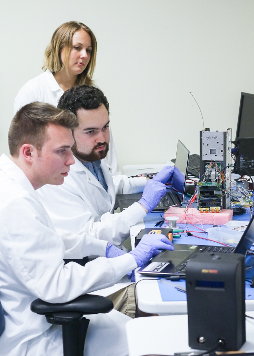 Photo of three scientists (1 female, 2 male) working on Q-PACE electronics at a desk.