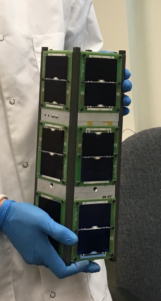 Photo of hands in blue latex gloves holding the Q-PACE satellite, a small device