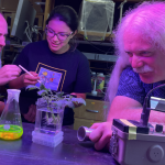 Photograph of three team members working at a table with a flask containing a yellow-green fluid, a clear canister with a rooted plant and a metal box.