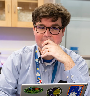 Portrait photo of a young man with short wavy brown hair and eyeglasses sitting at a laptop with his hand covering his mouth