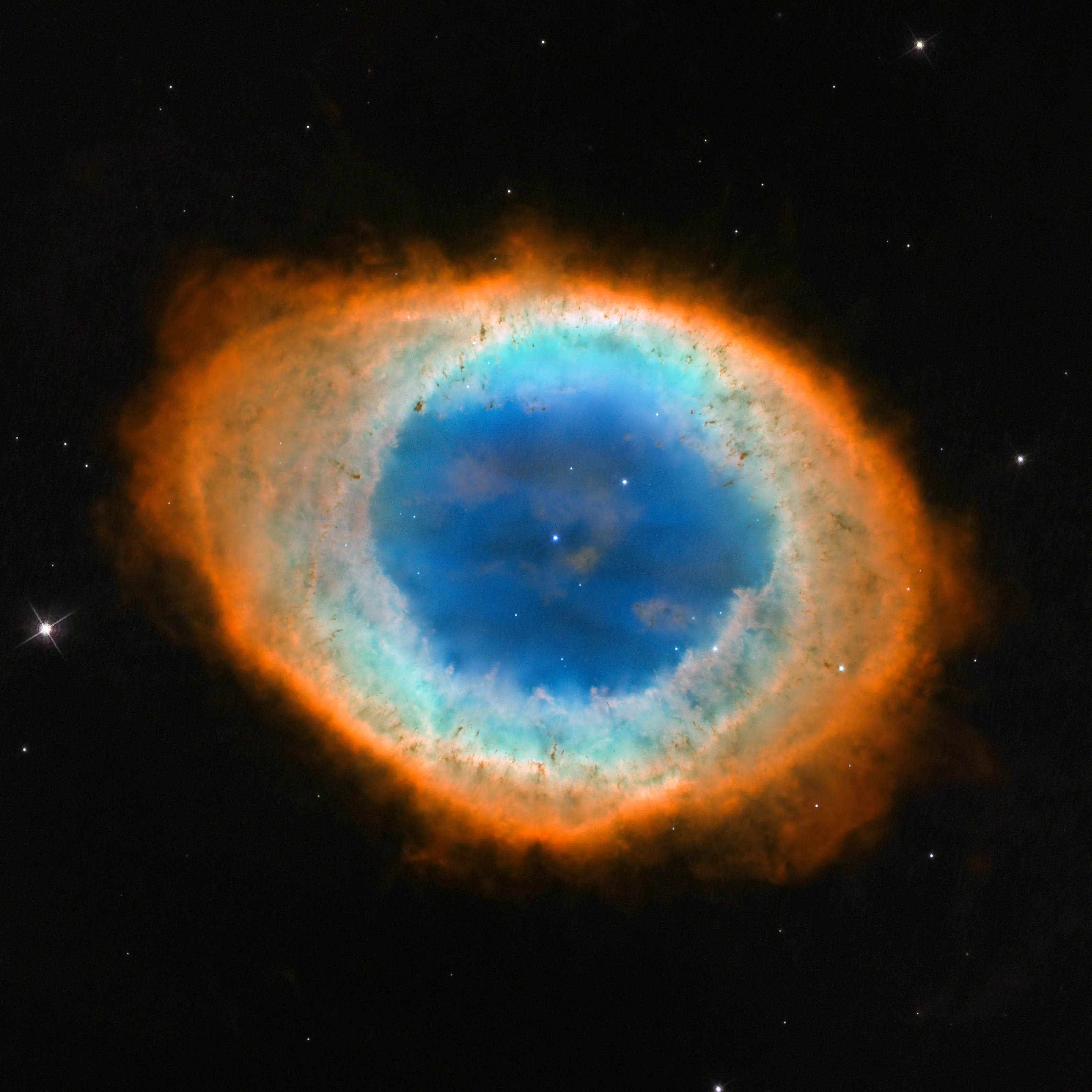 Black background holding an oval of light. The outer ring of the oval is orange-red. Moving inward the ring is more greenish-yellow. The center of the ring is bright blue with one small star at its center and another above and to the right of it.