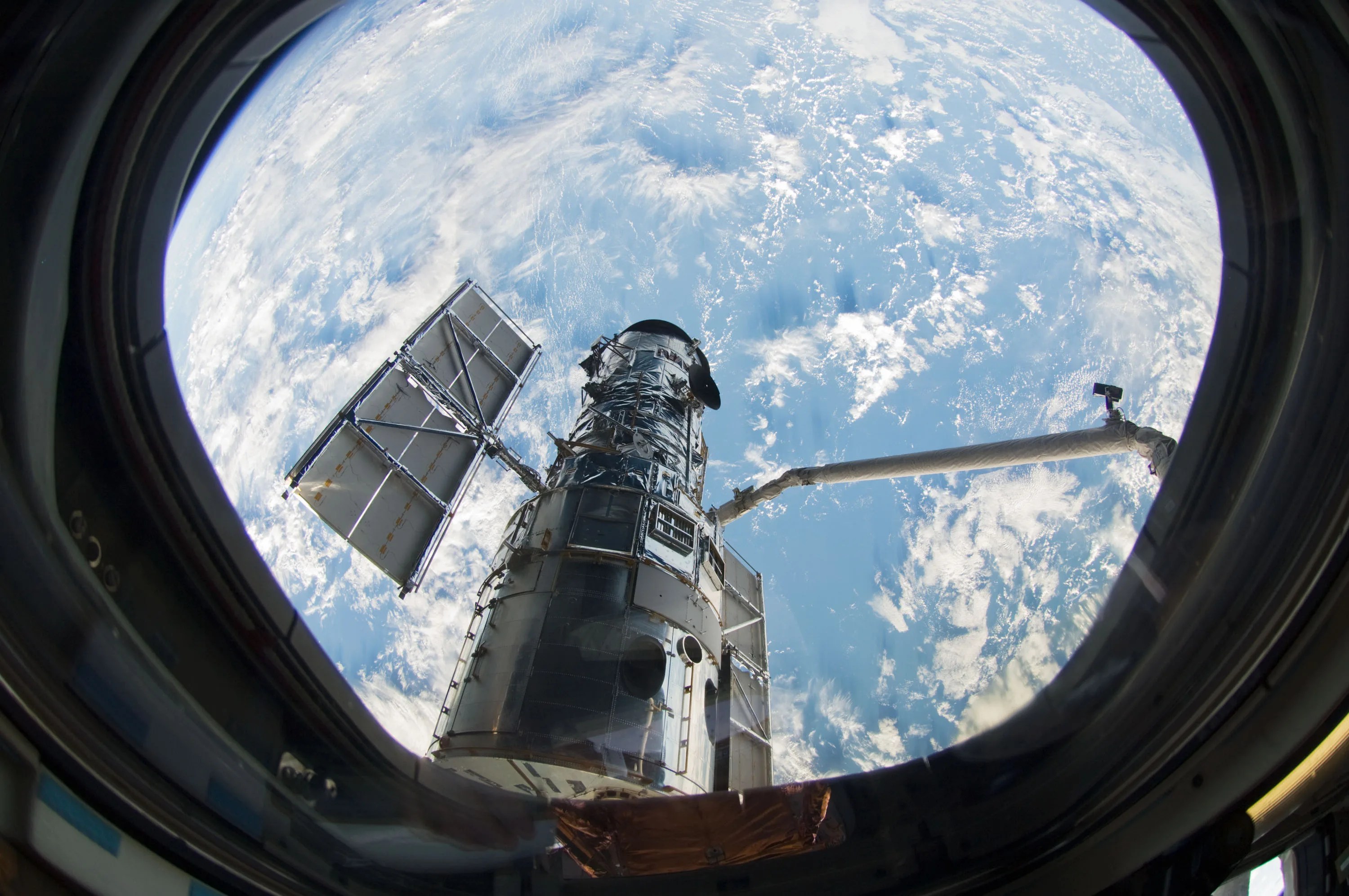 A window on the space shuttle Atlantis frames Hubble. One of Hubble's solar panels and the shuttle's robotic arm are visible through the window, and a blue, cloud-covered Earth fills the background behind the telescope.