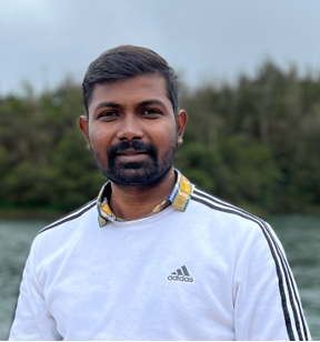 Portrait photo of a man with dark hair and dark facial hair wearing a white Adidas sweater; blurred trees are in the background