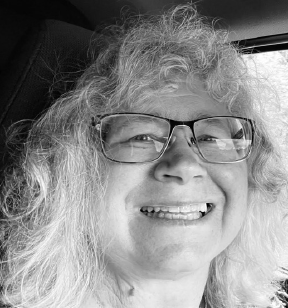 Black and white portrait photo of a smiling woman with grey hair and eye glasses and sitting in a car
