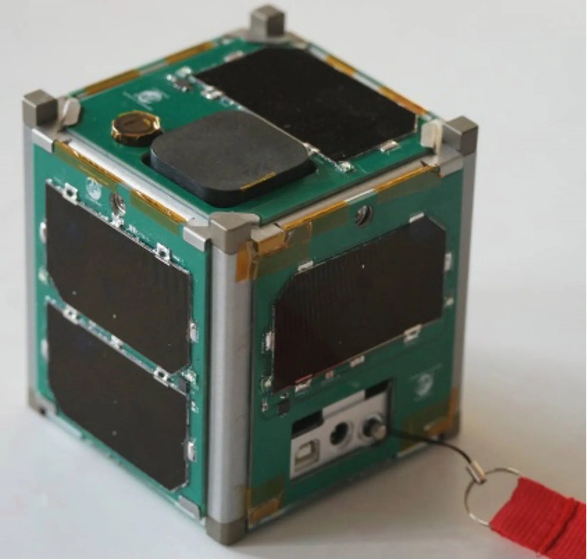 A small, green satellite rests on a white surface. It is in the shape of a cube with black rectangular panels that cover various spots on the outer surface.