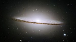 A bright hazy, oblate ball of stars fills the scene. Its core is bright white. Around the equator is a disk of bright stars and dark dust. The dust lane is prominent and bisects the sphere of stars.